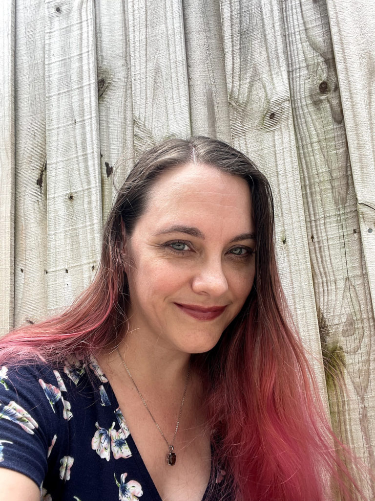 A photo of artist and writer Rebecca Wilcox. She is a white woman with gray eyes and hip-length brown hair dyed pink and wearing a navy blue dress and a smokey quartz pendant. A wooden fence. bleached pale by the sun, fills the background. Rebecca is smiling.